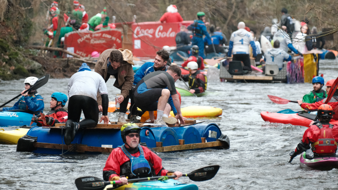 Kayakers can be seen on a river in between custom-made rafts with multiple people on top of them. The rafts are made from blue barrels bound together by wood.