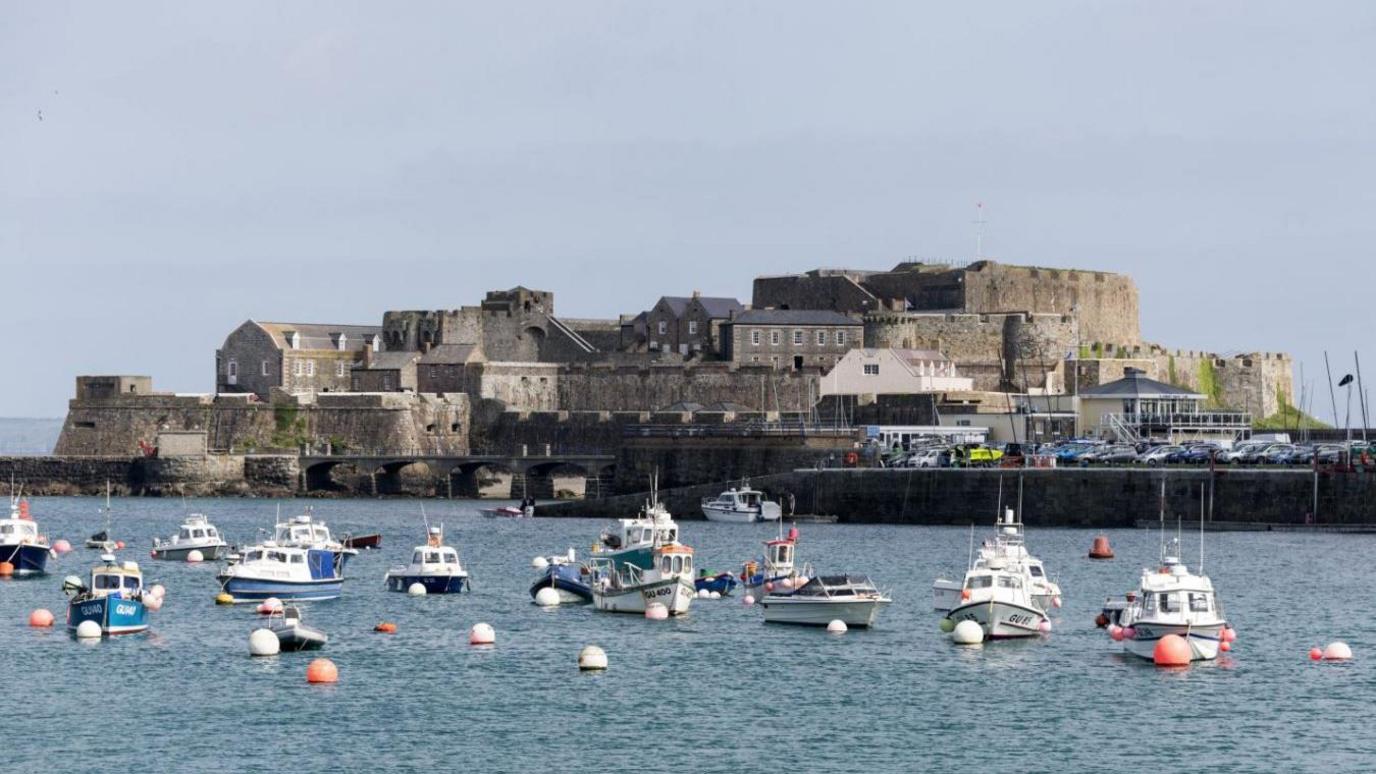 Castle Cornet, a large island castle on Guernsey that is largely surrounded by water. In the image there are several boats moored in front of the castle. There is also a full car park to the right of the structure. The day is largely clear with some light cloud coverage.