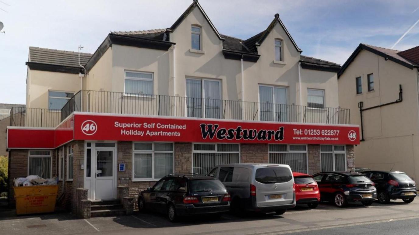 A detached building with two floors and dormer windows in the roof. A balcony with black metal railings rounds around the first floor. There is a large red sign below the balcony, with "Westward" in black letters and white writing describing the type of business and contact details. Five cars are parked in bays in front of the building