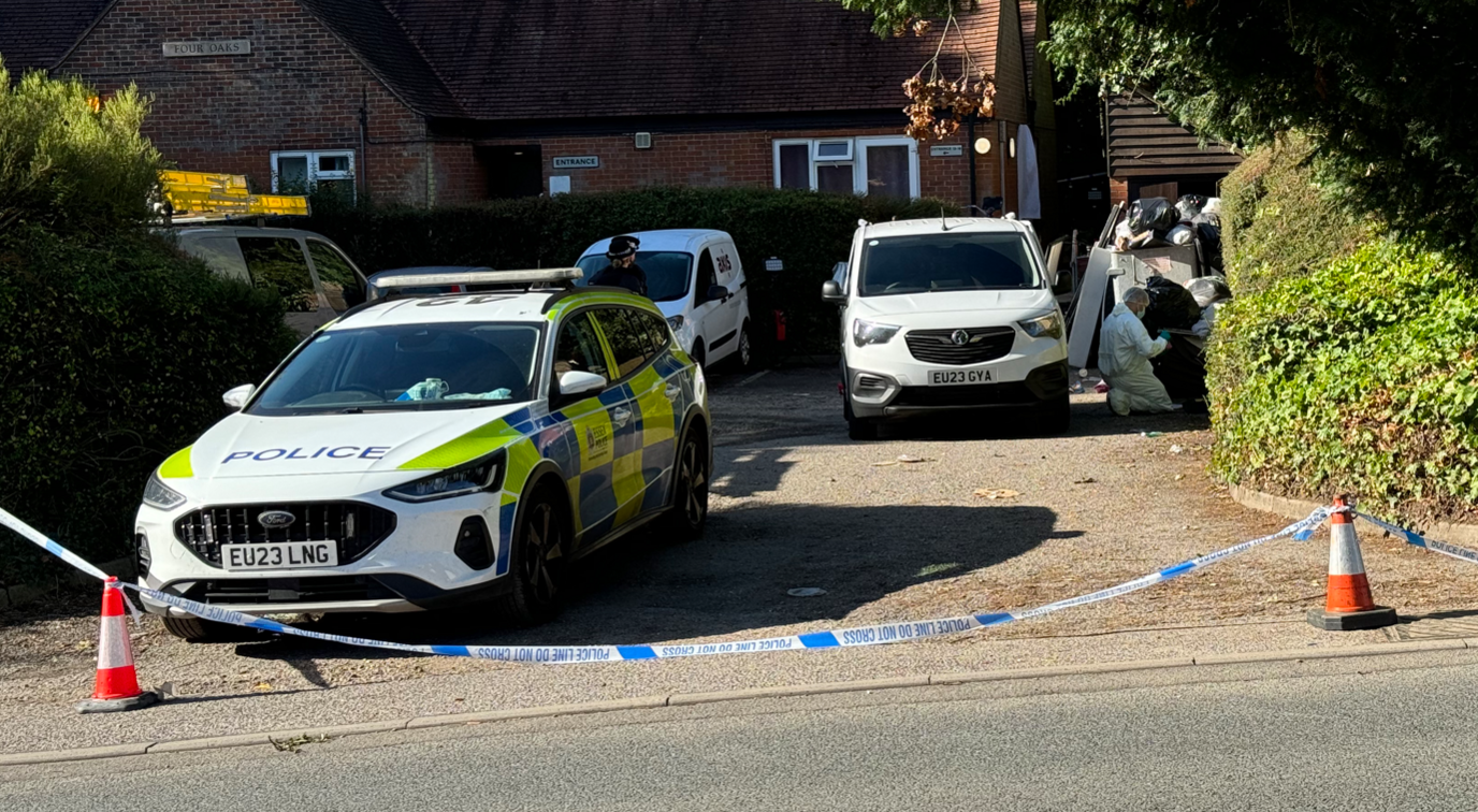 A police car and forensics vans parked on a driveway. Police tape blocks entrance to the property.