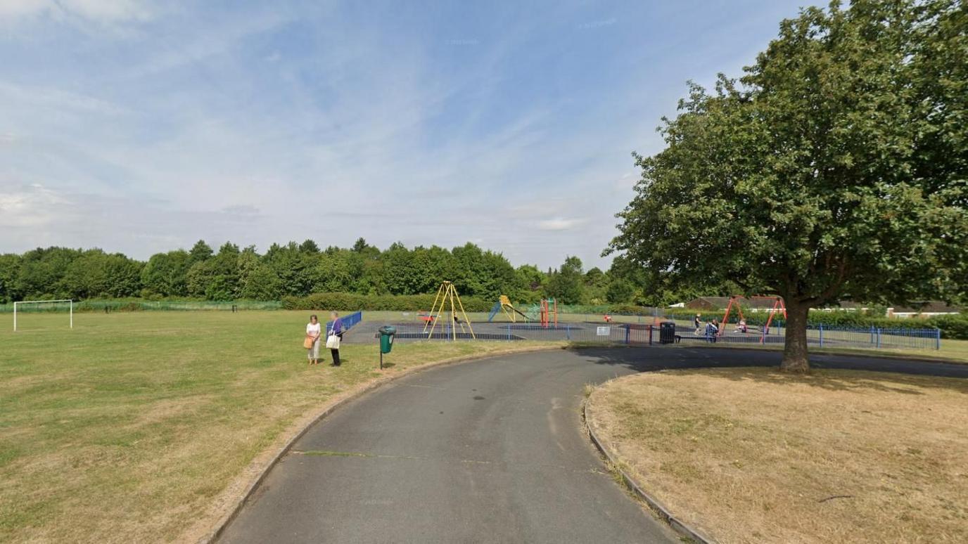 An open green space under blue skies. There is a set of goalposts and a path leads to a small playground where children are playing on the swings. Two people stand in the foreground talking. Trees are visible in the distance and on a smaller patch of grass near the playground.