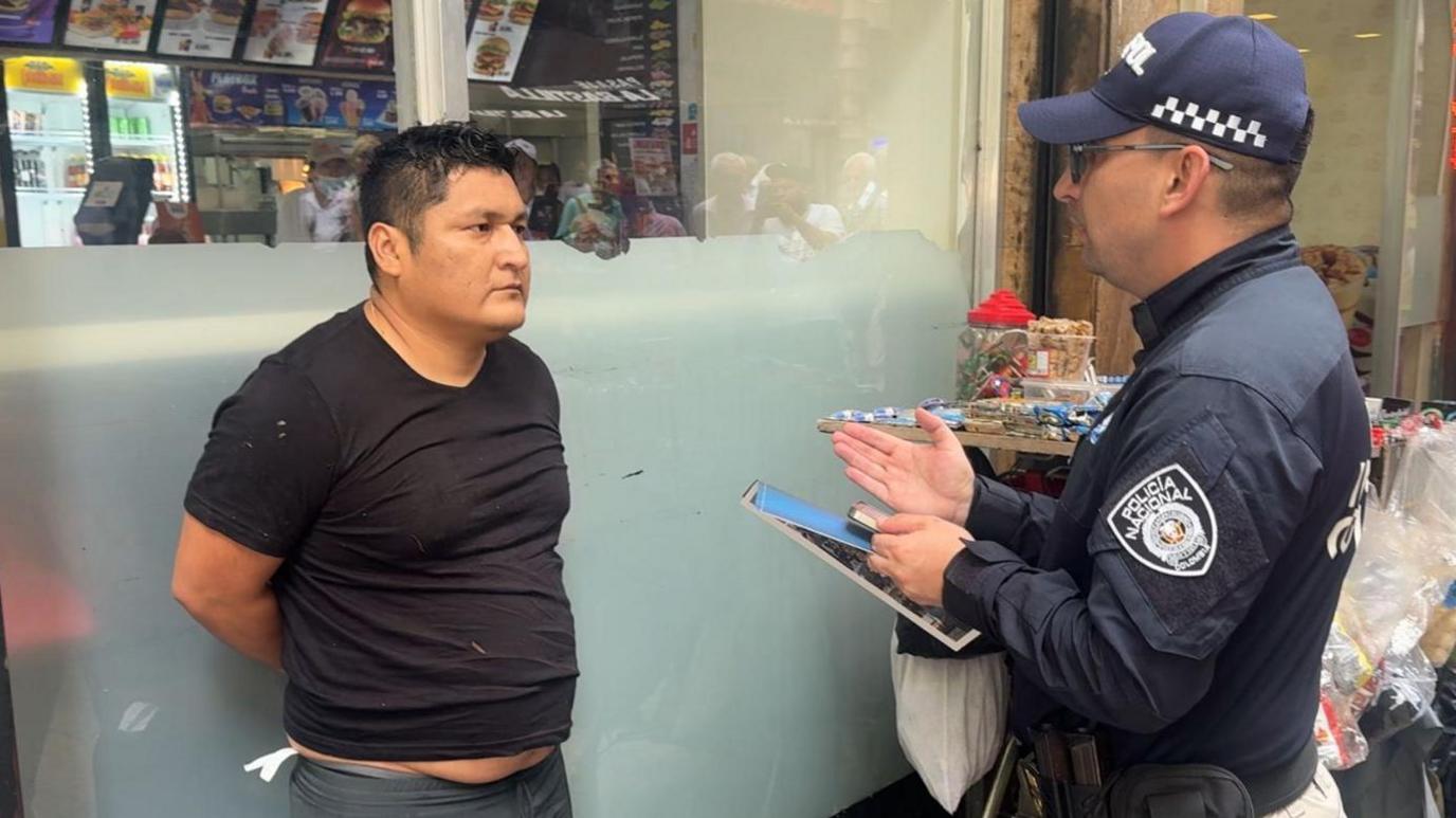 A police officer speaks to a man in a black T-shirt who has his hands behind his back after being arrested.
