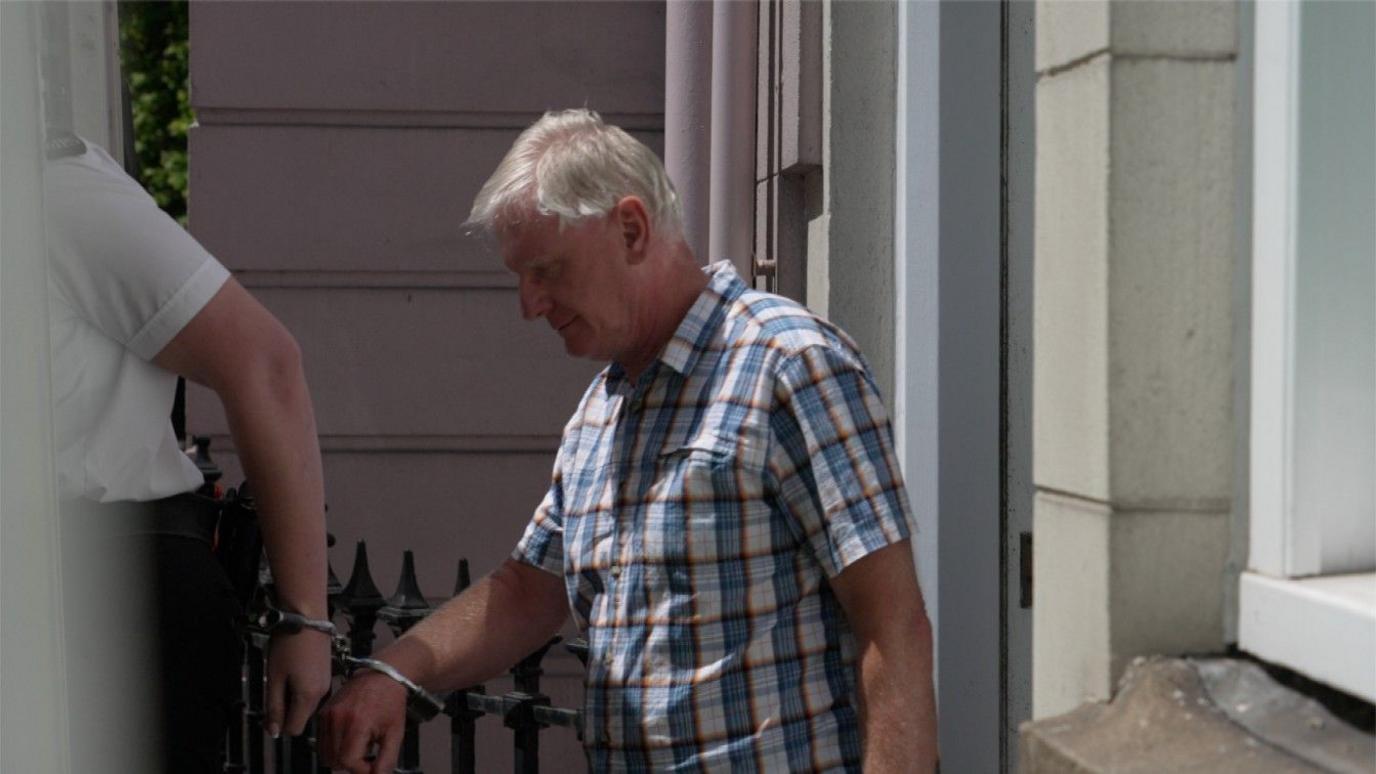 Piotr Antoni Glas in a checkered shirt and dark pants standing between a pinkish building and a large white police vehicle. In the foreground, there's an iron fence with pointed tops. Another person is partially visible behind the truck's side mirror, handcuffed to Glass leading him into the van.