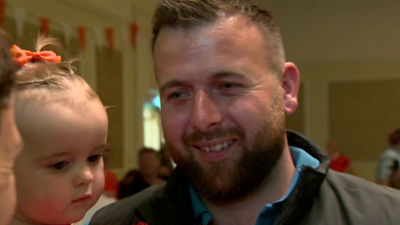 A man holds his baby. He's wearing black and blue. The baby has an orange bow in her hair. 