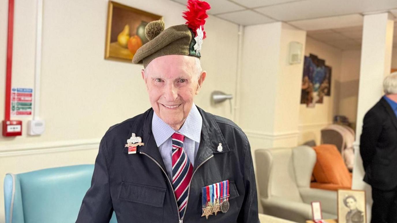 Dennis Smith stands, smiling, in his care home, with military medals pinned to his dark, zip-up jacket. His green beret has a red hackle. Three medals are pinned to his chest and there is a badge on each lapel.