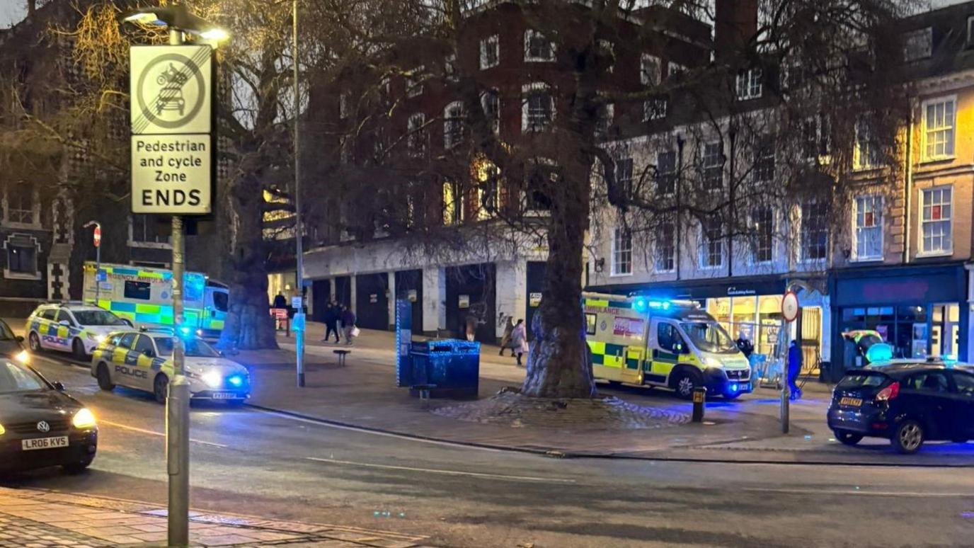 Two police cars and two ambulances in a city centre street in late afternoon.There are trees in the street and shops.