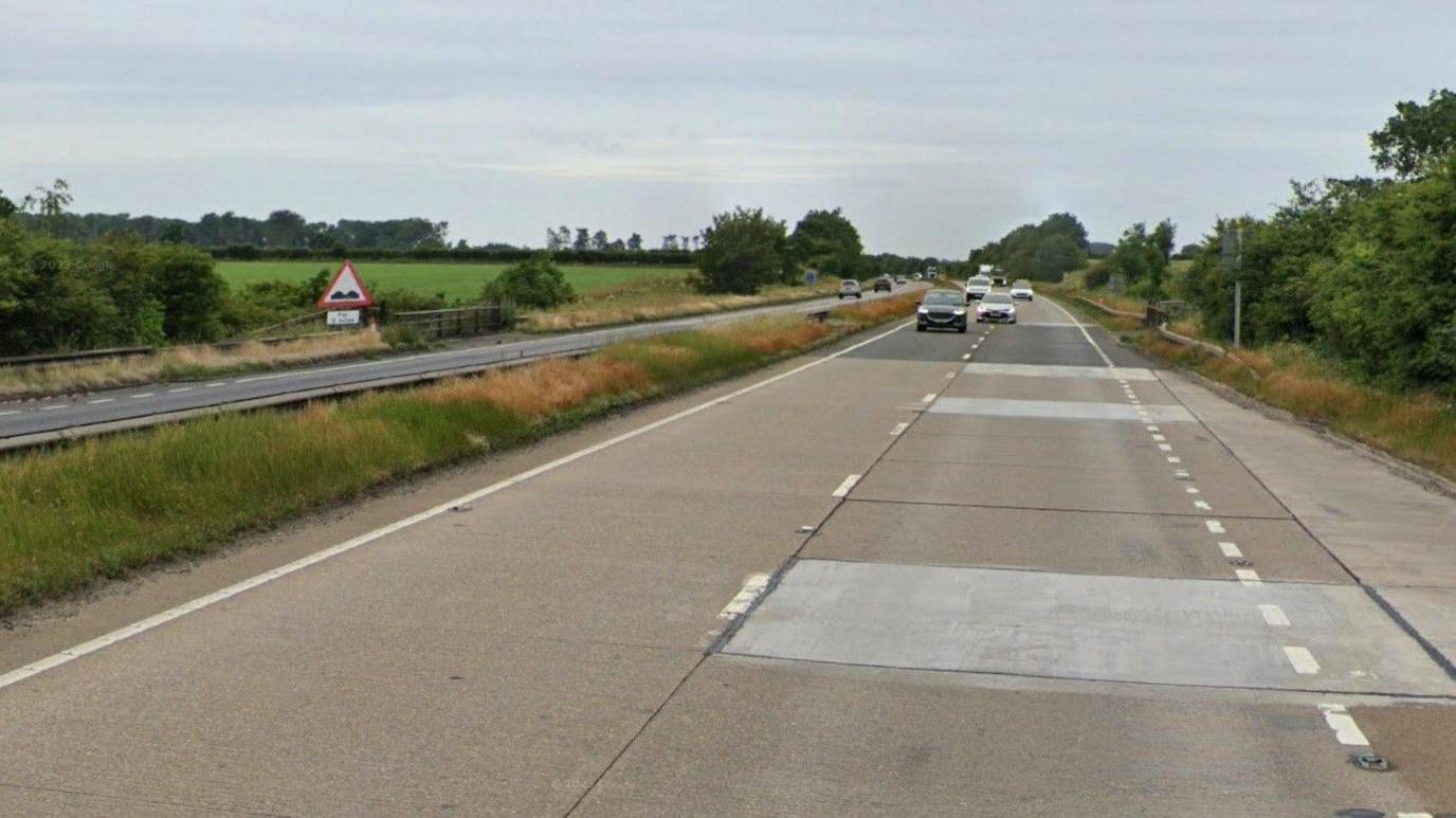 The A180 dual-carriageway, with a surface made up of patched concrete sections, and a grassed central reservation. Cars can be seen in the background, travelling towards the camera. To the left are hedgerows and green fields