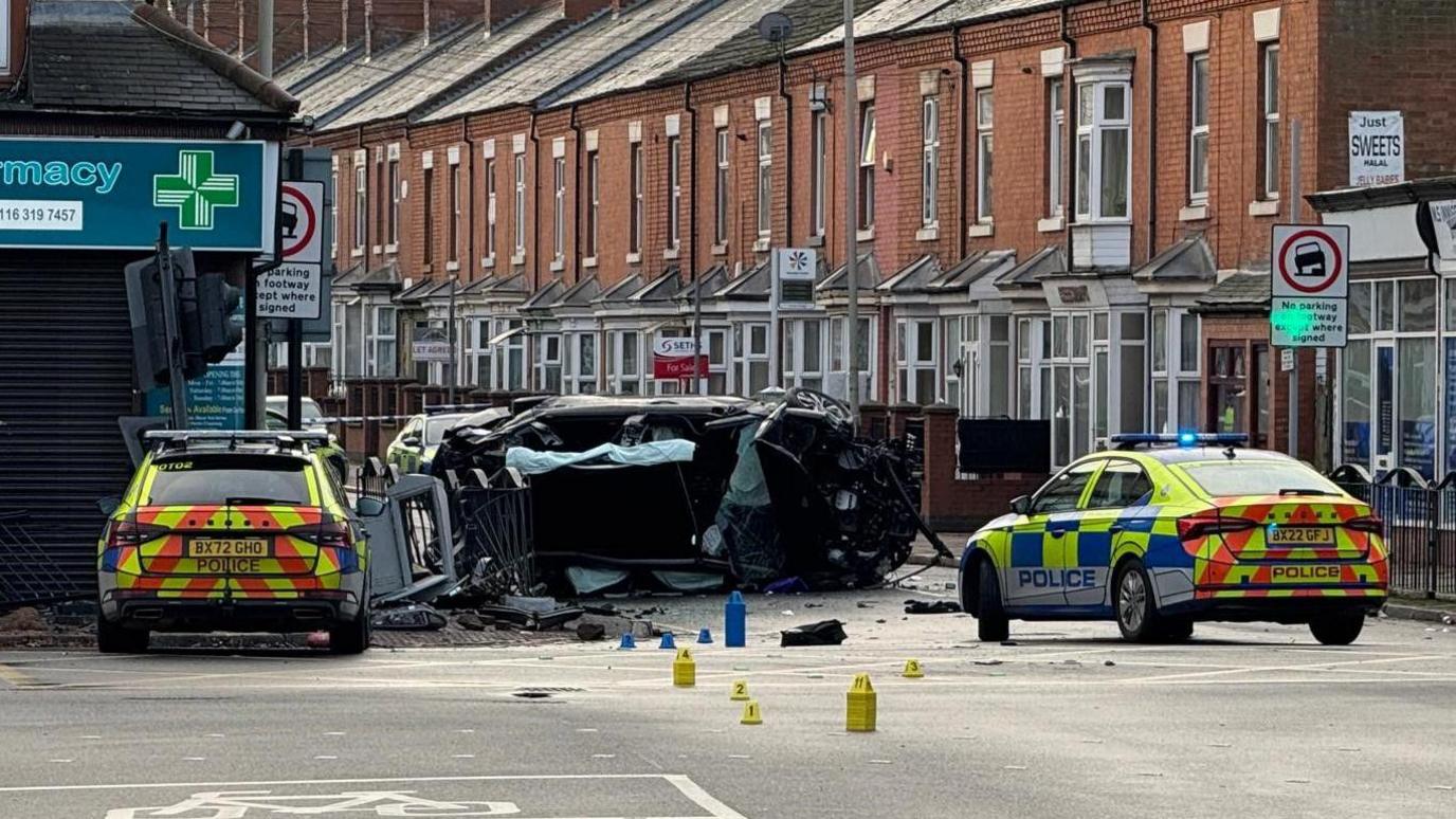 Two police cars at a junction. Two damaged cars, one flipped on its side can be seen, with debris in the road