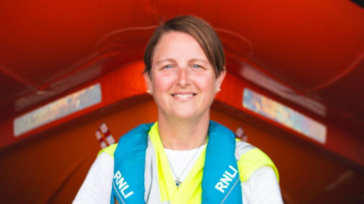 A portrait photo of Sonia Boydell smiling at the camera. She is wearing a grey long-sleeve top with a blue vest with RNLI written on it. She has short straight brown hair swept to the side and blue eyes.
