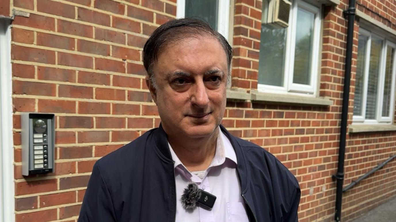 A man stands outside a block of flats. He has dark hair with grey sides. He wears a blue jacket and a white shirt. An interview microphone can be seen attached to his shirt.