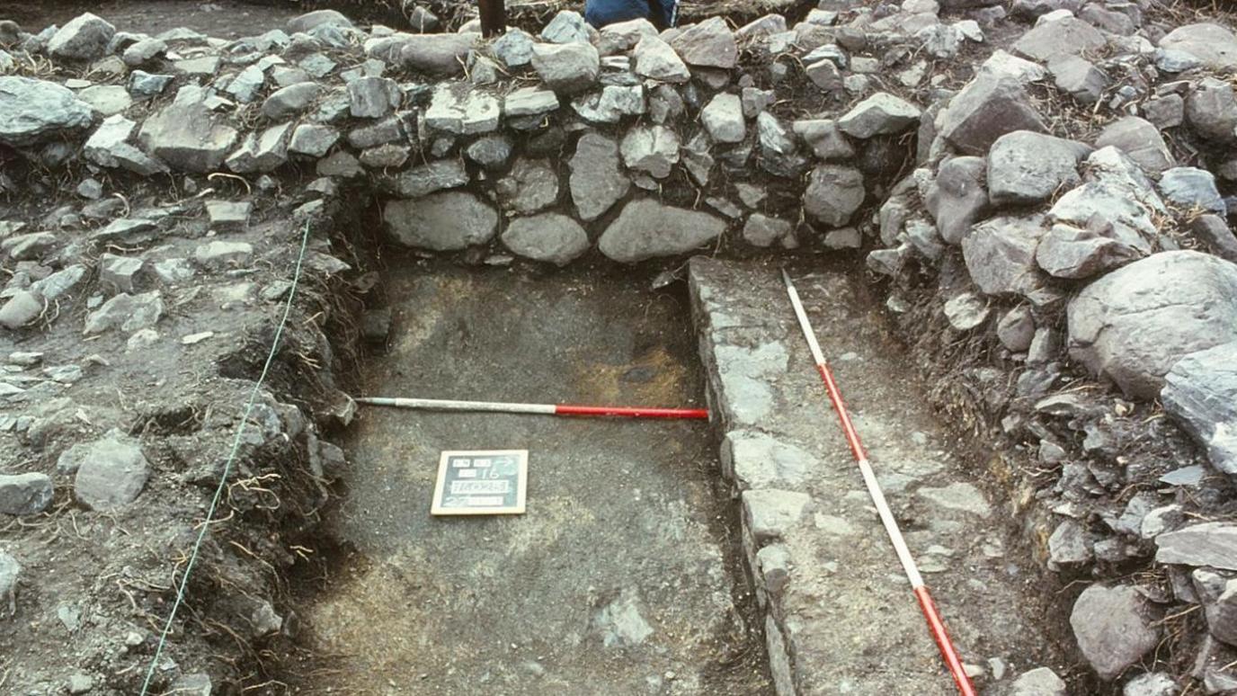 An exposed area of stone-built walls and a floor. There are red and white measuring poles laid out on the ground. 