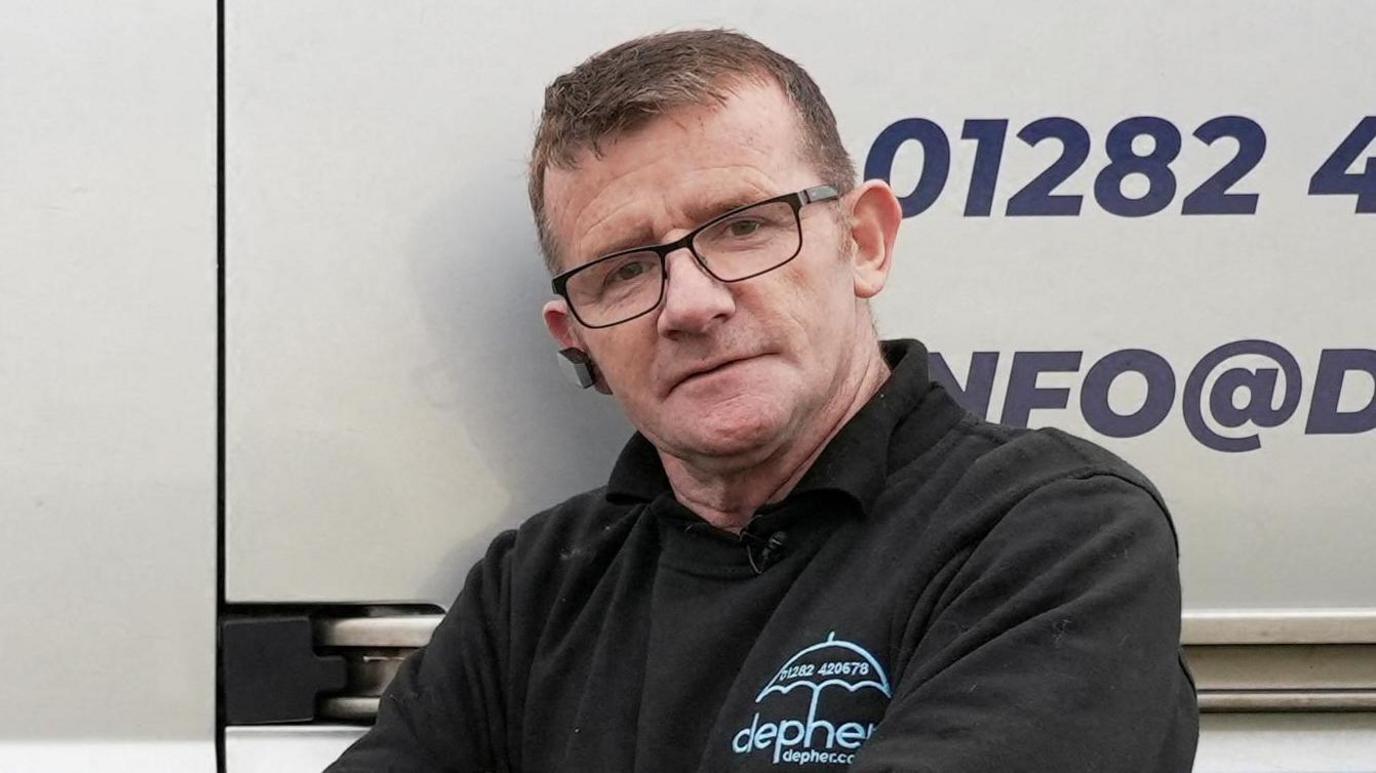 James Anderson standing in front of his van. He is wearing a black jumper with branding for his company Defer. He has short brown hair and black glasses.