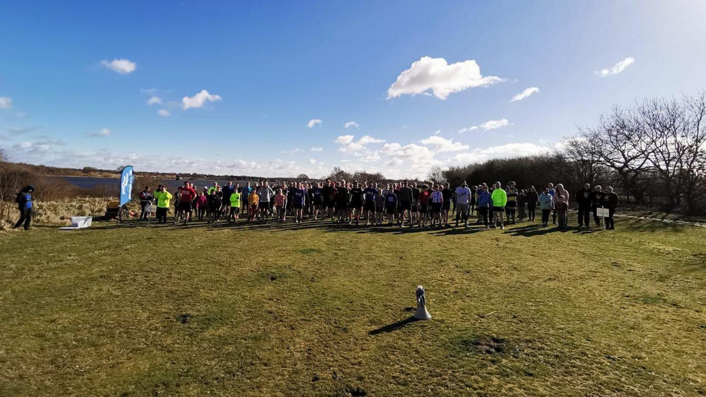 About 50 runners line up at Alton Water at the start of a 5km run. The reservoir is visible in the background.