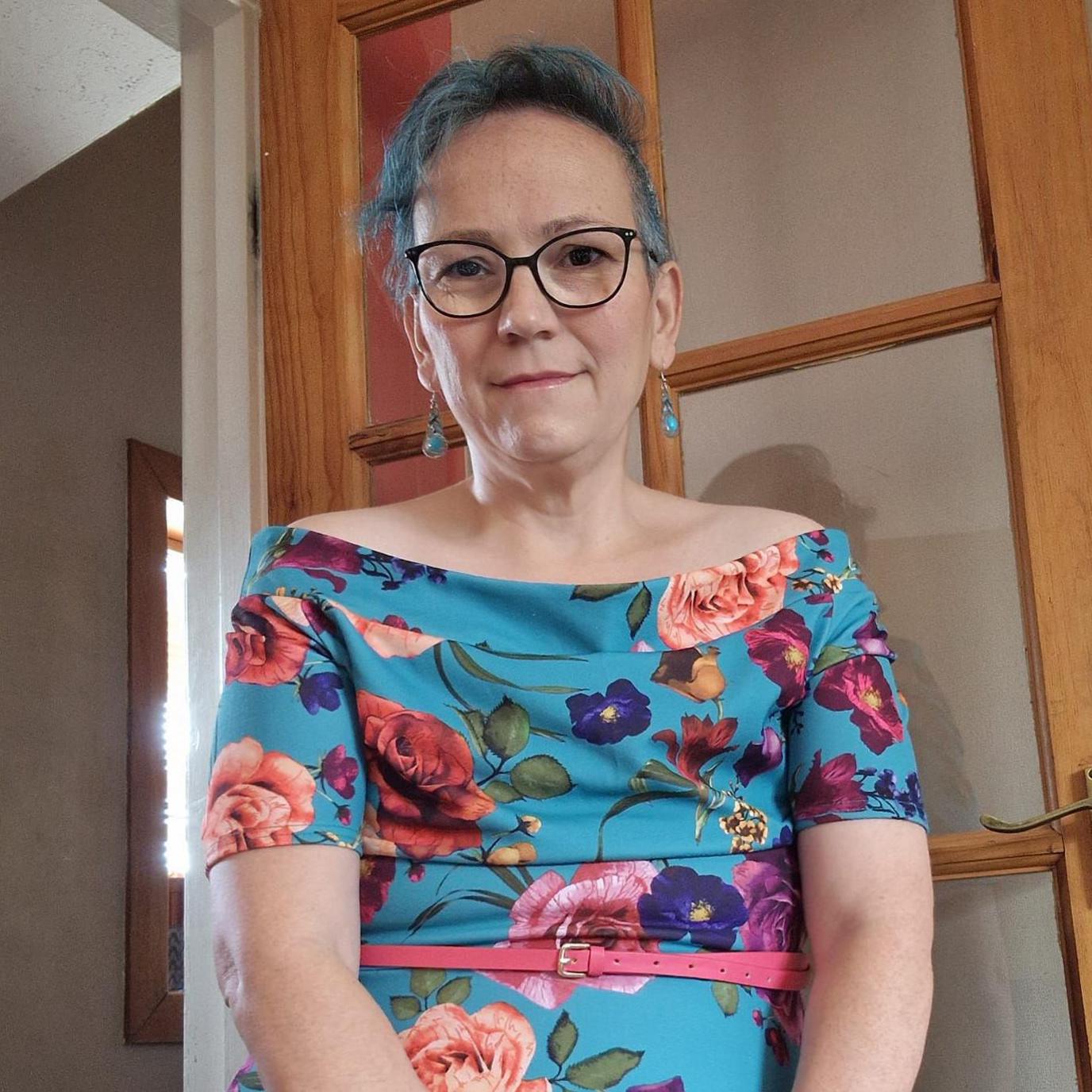 Jenni Christieson is standing with her hands clasped in front of her. She is wearing a blue floral dress with a slim pink belt around the waist. She is wearing glasses and her short hair is dyed blue