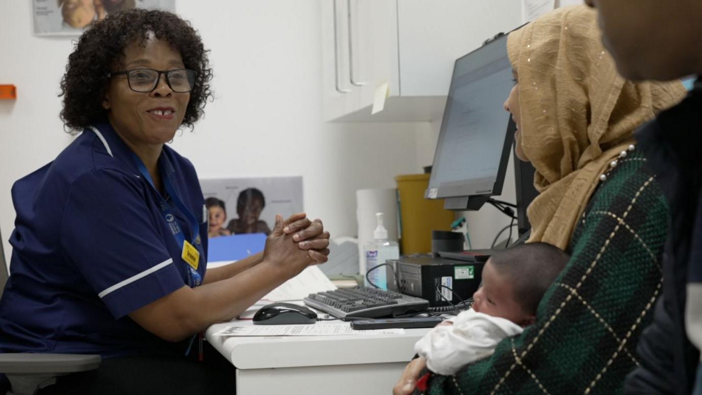 Midwife Irene Namugere is seen sitting at a desk in a clinical setting, speaking with a mother and baby. Irene is wearing a navy-blue uniform with white trim, glasses, and a name badge. The mother, partially visible, holds her baby while seated across from Irene. 