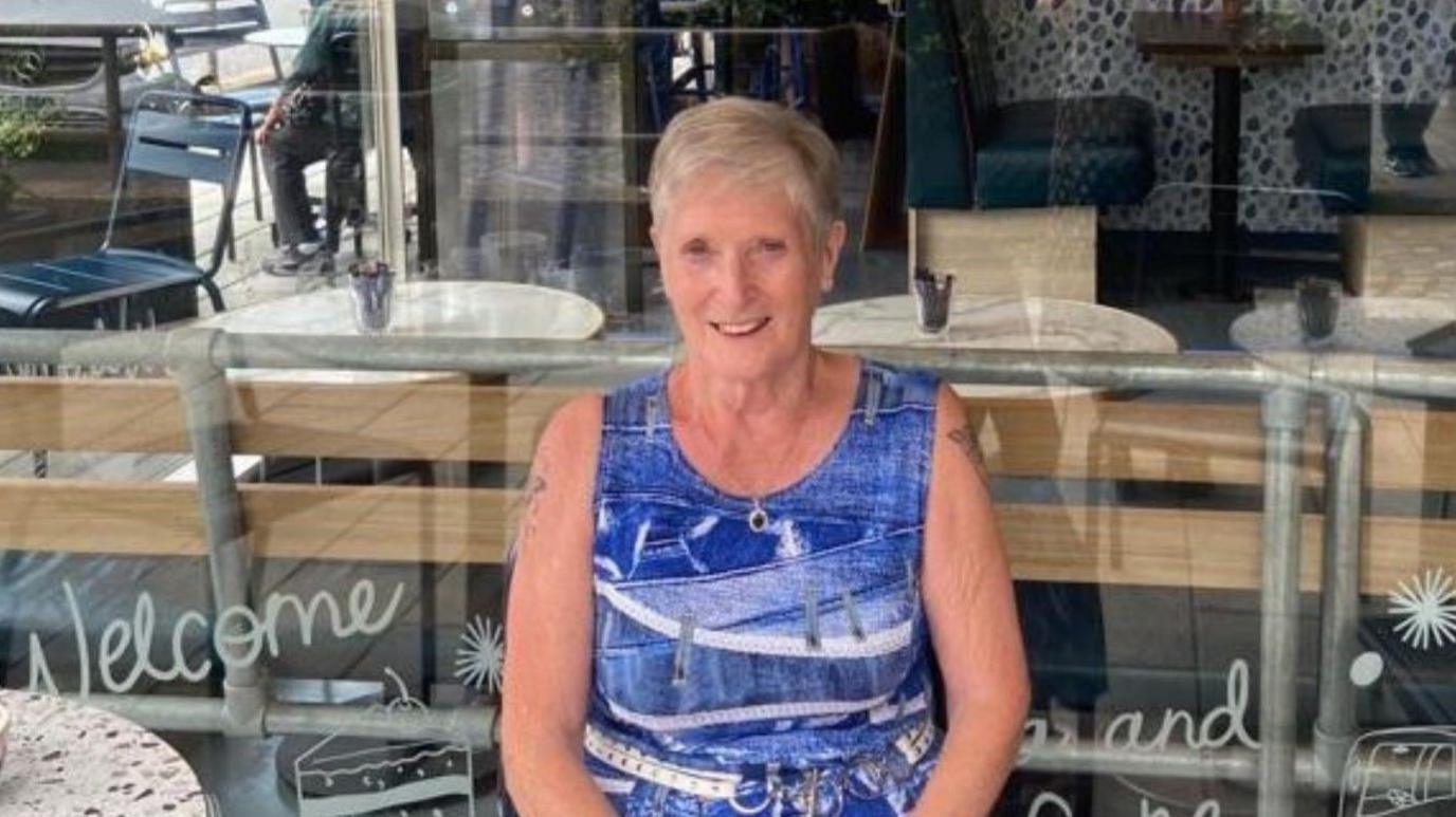 A woman in a blue dress sits outside a cafe smiling directly at the camera