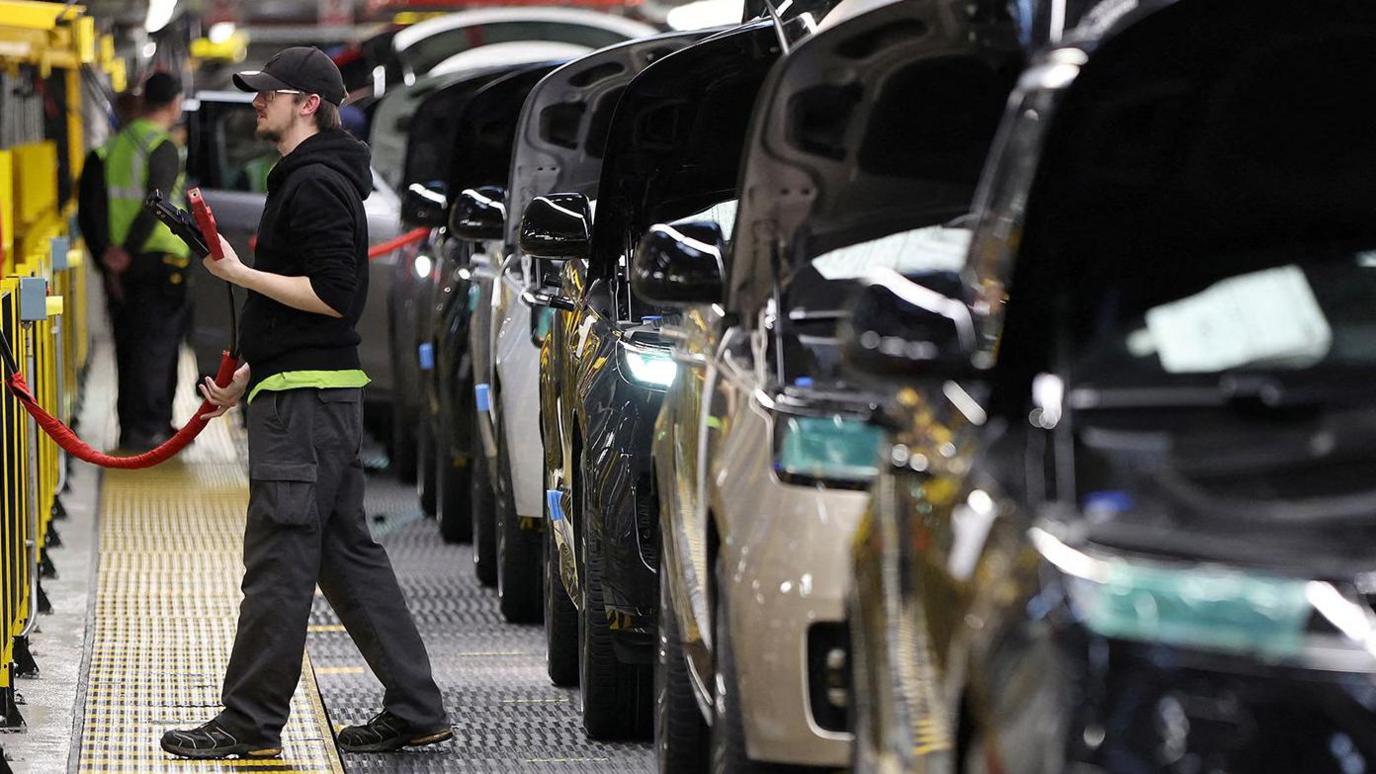 An assembly line in a car manufacturing plant with multiple cars lined up behind one another, hoods open. A worker in the foreground holds a red tool connected to a hose, while another worker is visible further down the line. The industrial setting includes various equipment surrounding the assembly line.