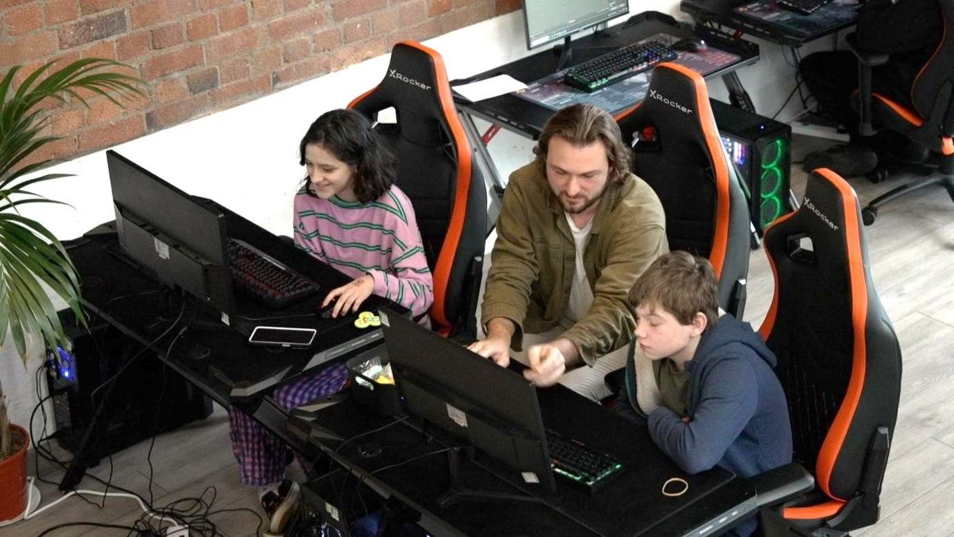 Two students at modern-looking computer setups, sitting in gaming chairs, with a tutor sitting between them. The computers and desks are black. The tutor is a man in a white t shirt with an open olive shirt over the top. He is showing the pupils something on the screen. The girls is in a pink top with green stripes, the boy is in a navy hoody.