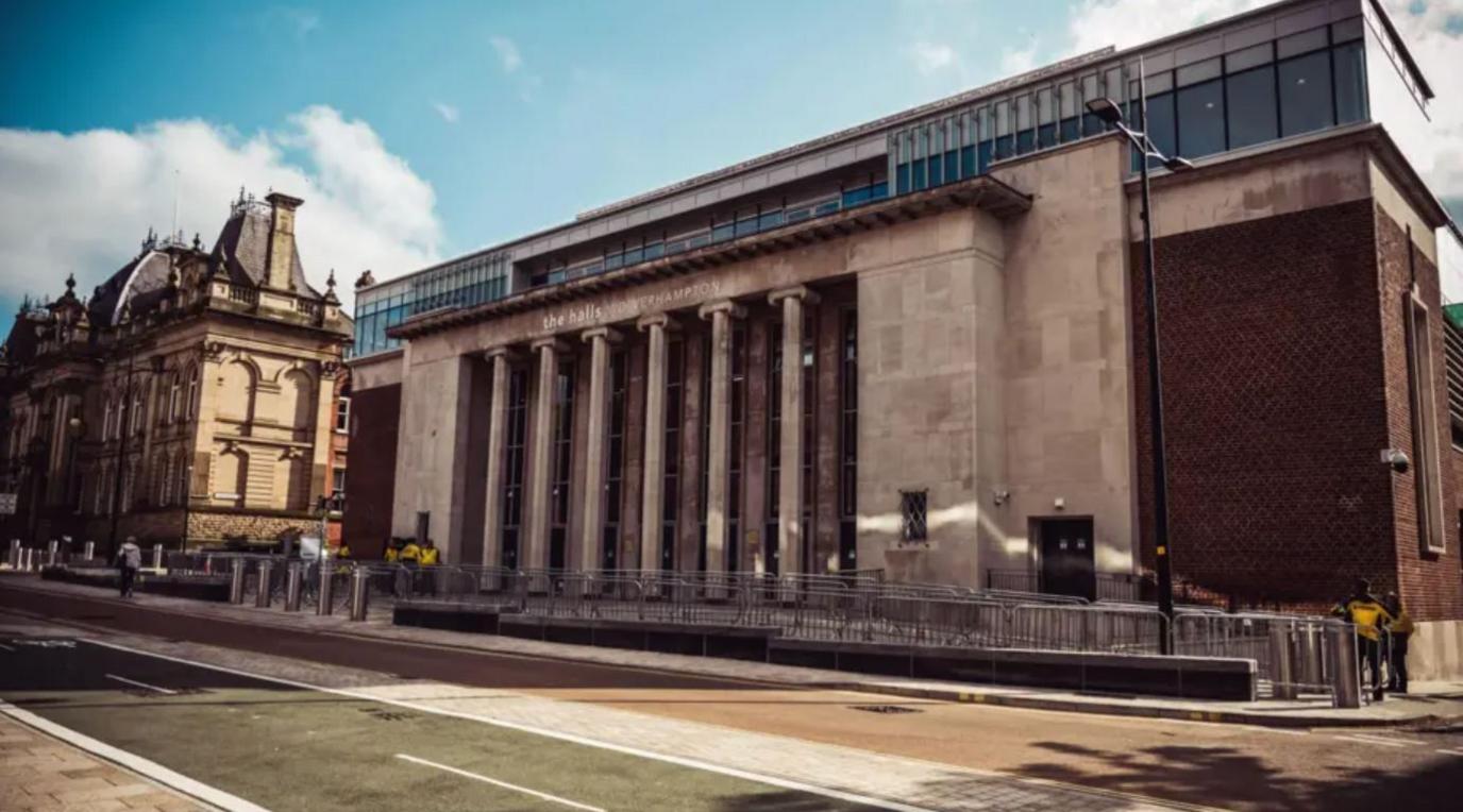 An external shot of University of Wolverhampton at the Halls from the opposite kerb. It is a tall stone building with six stone columns flanking the front. A roof extension is more modern, with windows the full way along. Staff in yellow jackets stand by metal railings which have been set up outside.