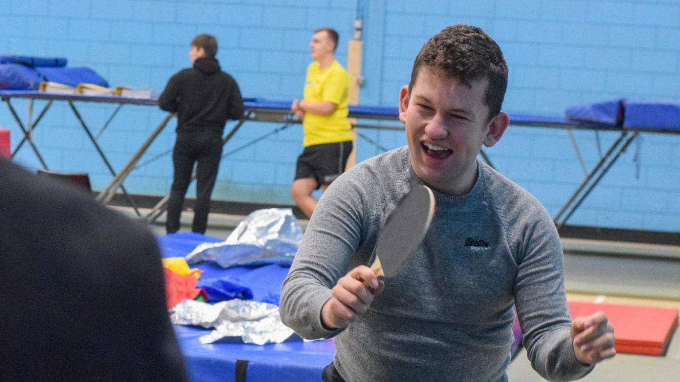 A smiling young man holds a table tennis bat, ready to receive the ball, in a sports hall. He has short, brown hair and wears a grey, long-sleeved sports shirt. Behind him, crash mats can be seen, while two other participants are standing in front of a bright blue brick wall.