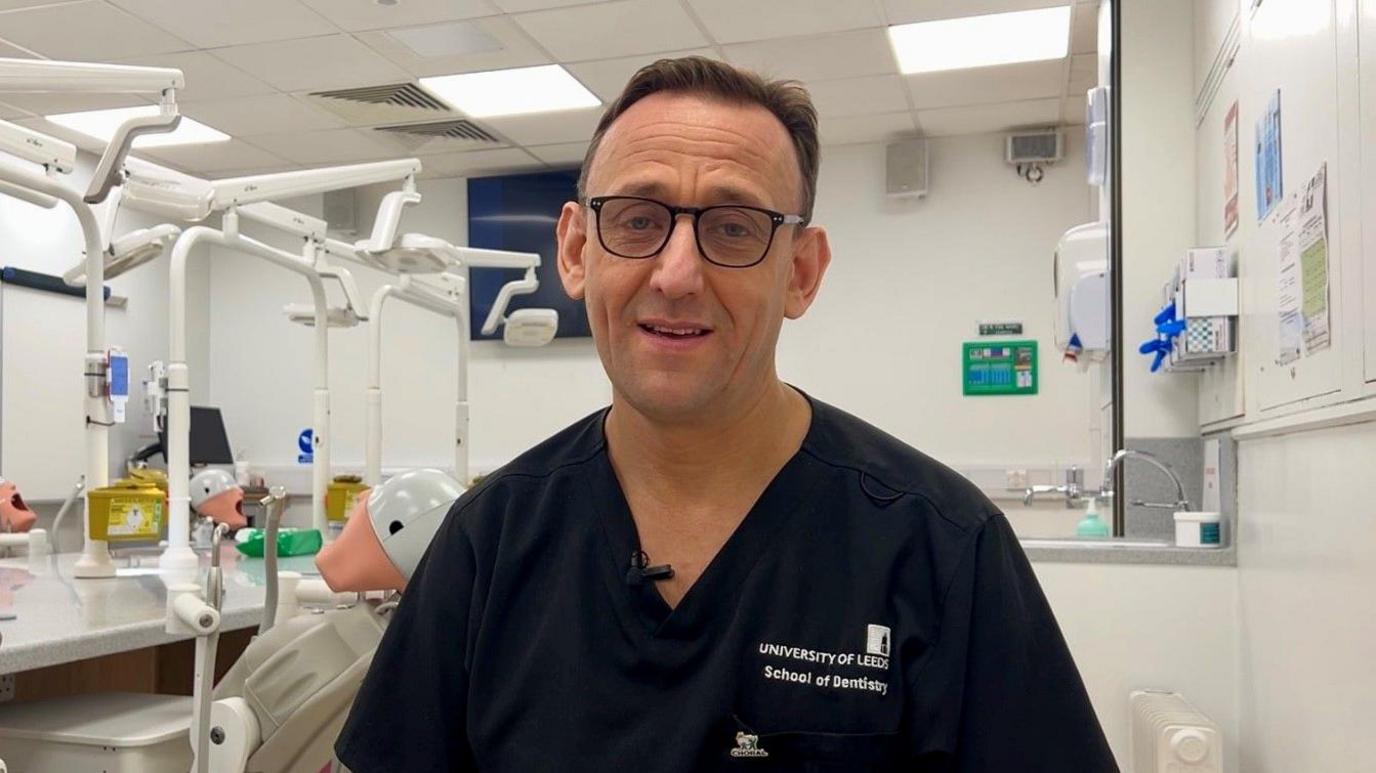 A man in his fifties, wearing blue surgical scrubs, is standing in a clinical setting that is used to train dentists. Behind him is a row of desks with mannequins that are used by student dentists to practise their skills on.