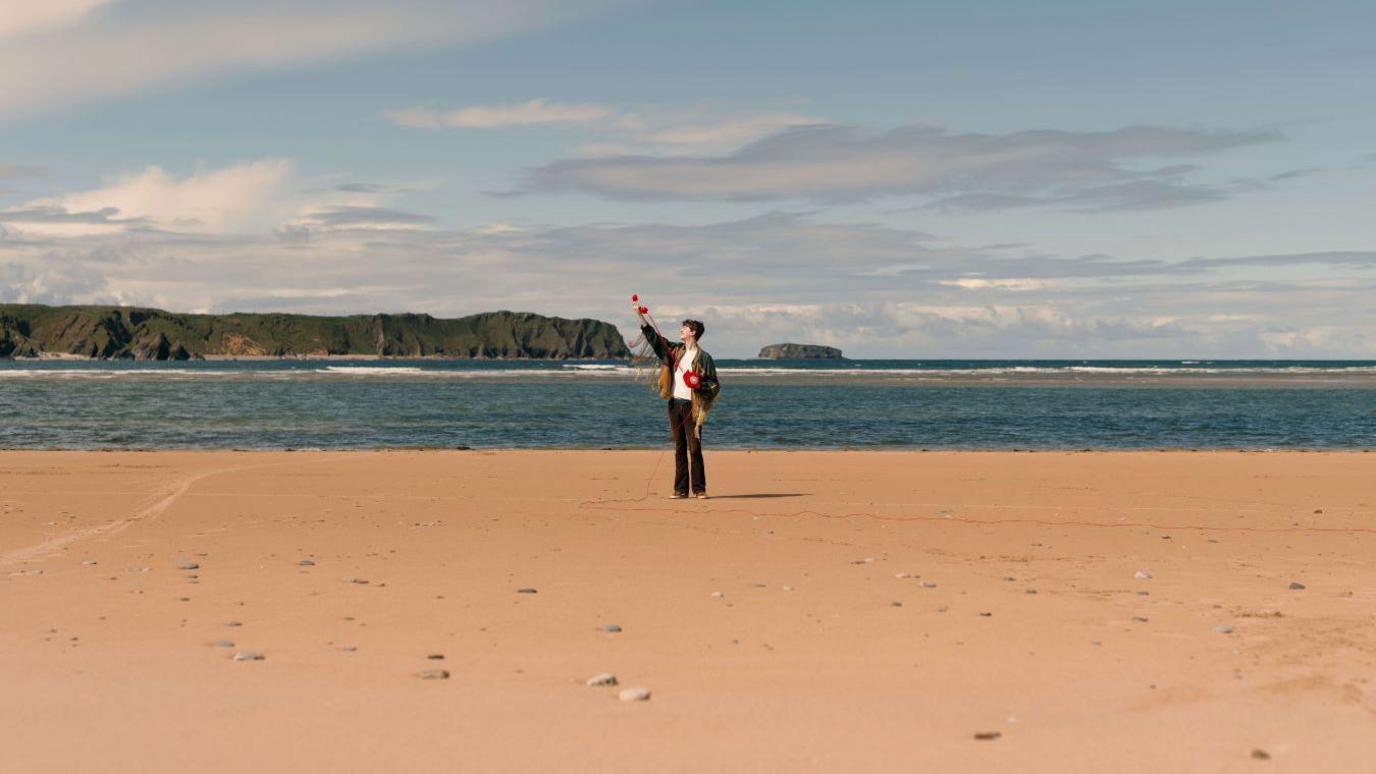 A man wearing a blue jacket stands on the beach. He is far away from the camera. It is a sunny day, and the sea is visible behind the man.