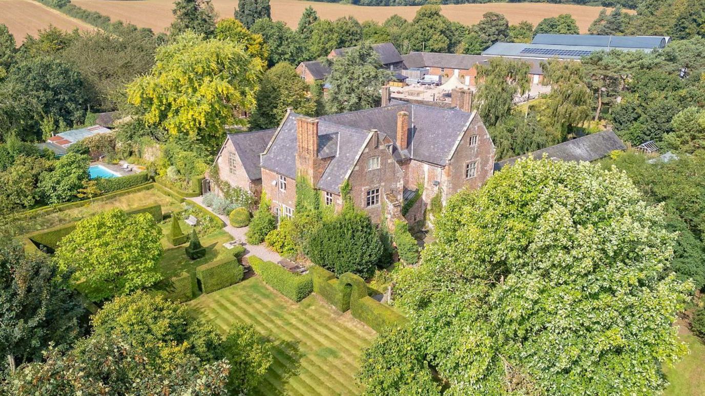 An aerial view of a large Elizabethan house, surrounded by green fields and woodland