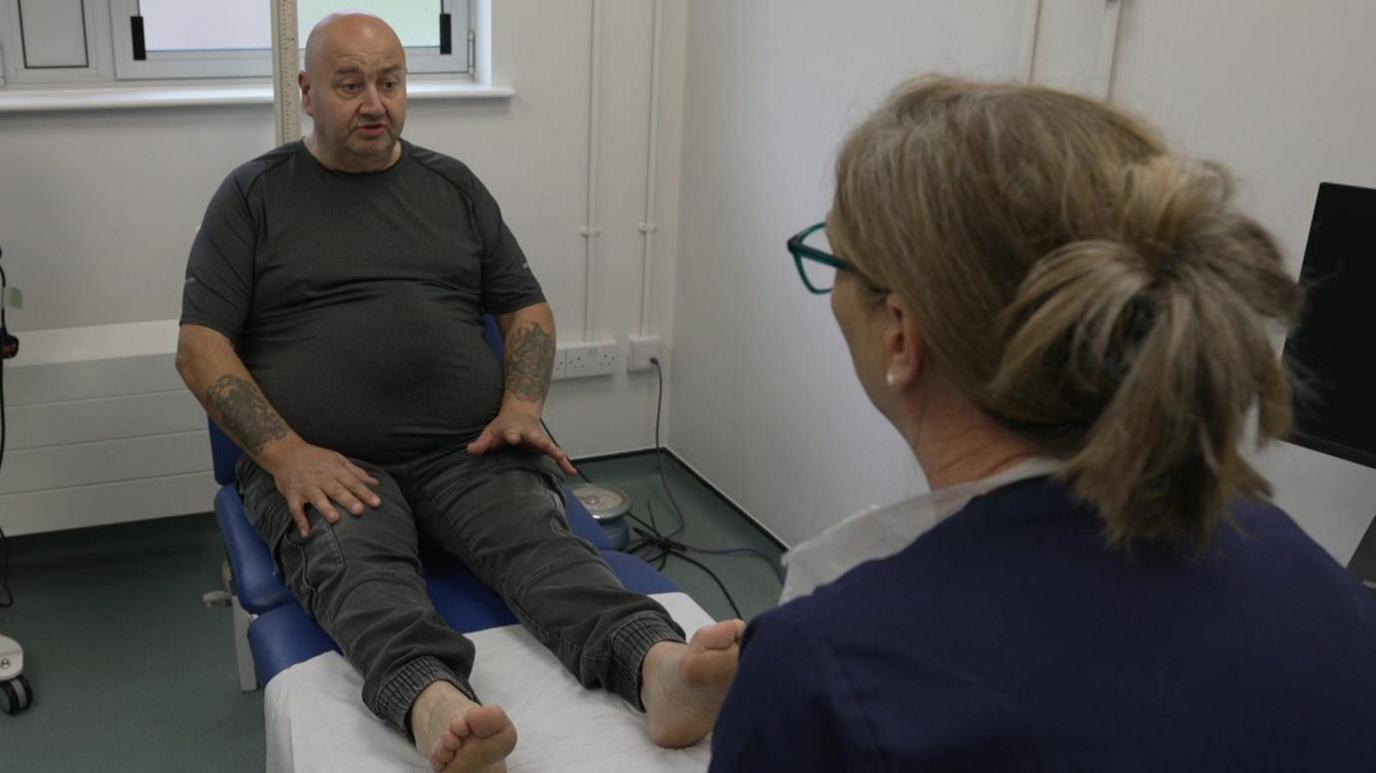 The picture shows a person is sitting on a medical examination table with their shoes and socks off. A healthcare professional, is seated nearby. The room includes medical equipment.