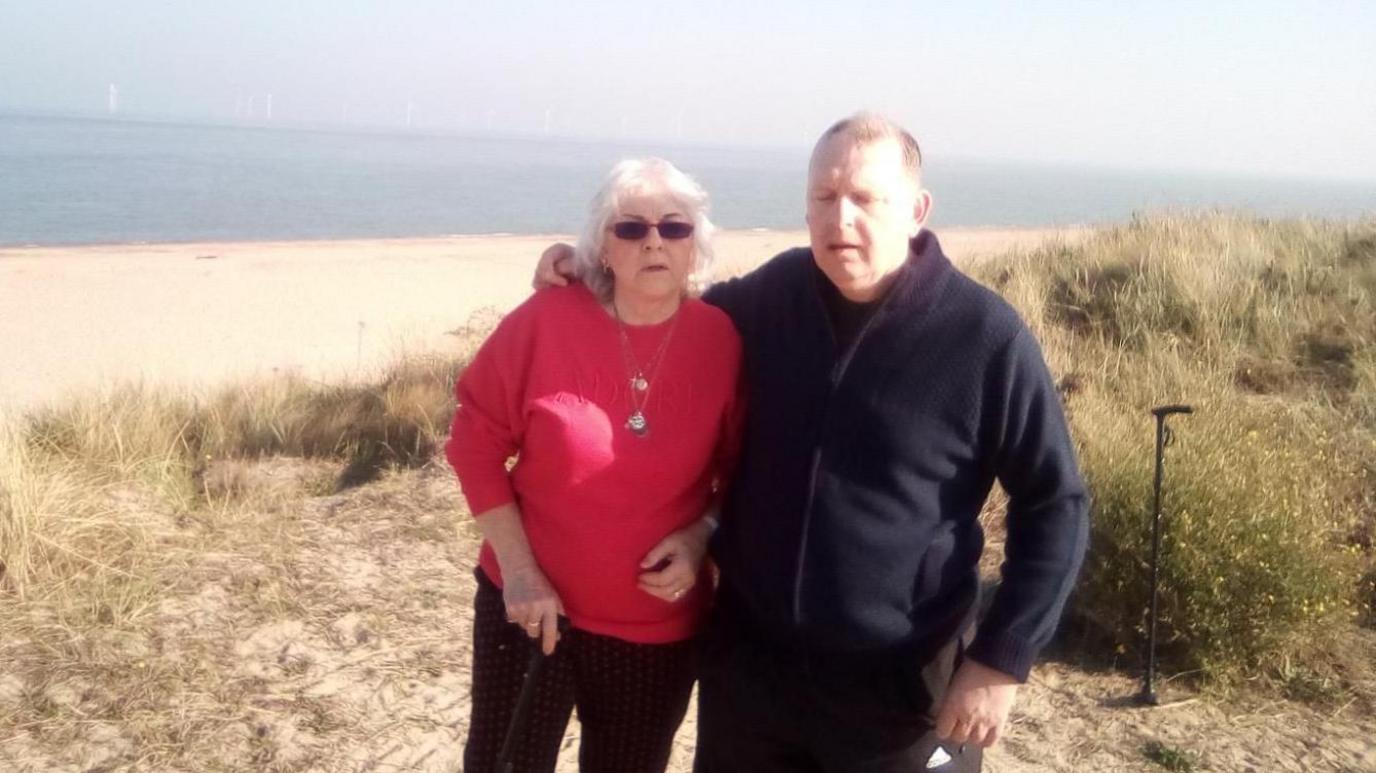 Barbara and her adult son Andy are stood on a beach 