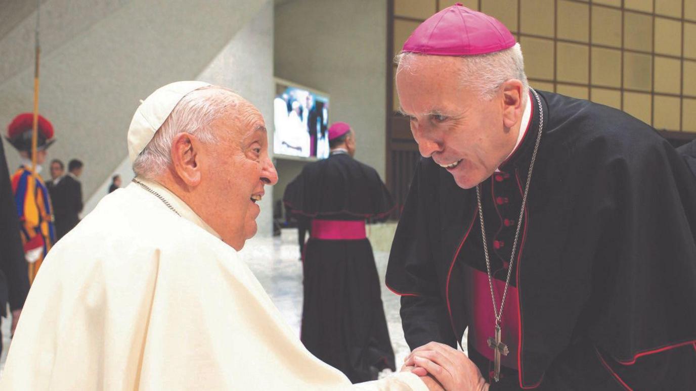 Pope Francis and Catholic Bishop of East Anglia Peter Collins shaking hands. Pope Francis is wearing all white whereas Bishop Peter is wearing black and pink.