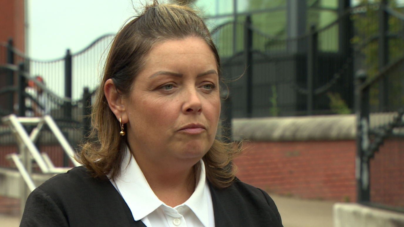 Ms Hargey is wearing a white blouse and a black jacket. Her brown hair is half up and half down. She has gold hoop earrings on. There are railings in the background.