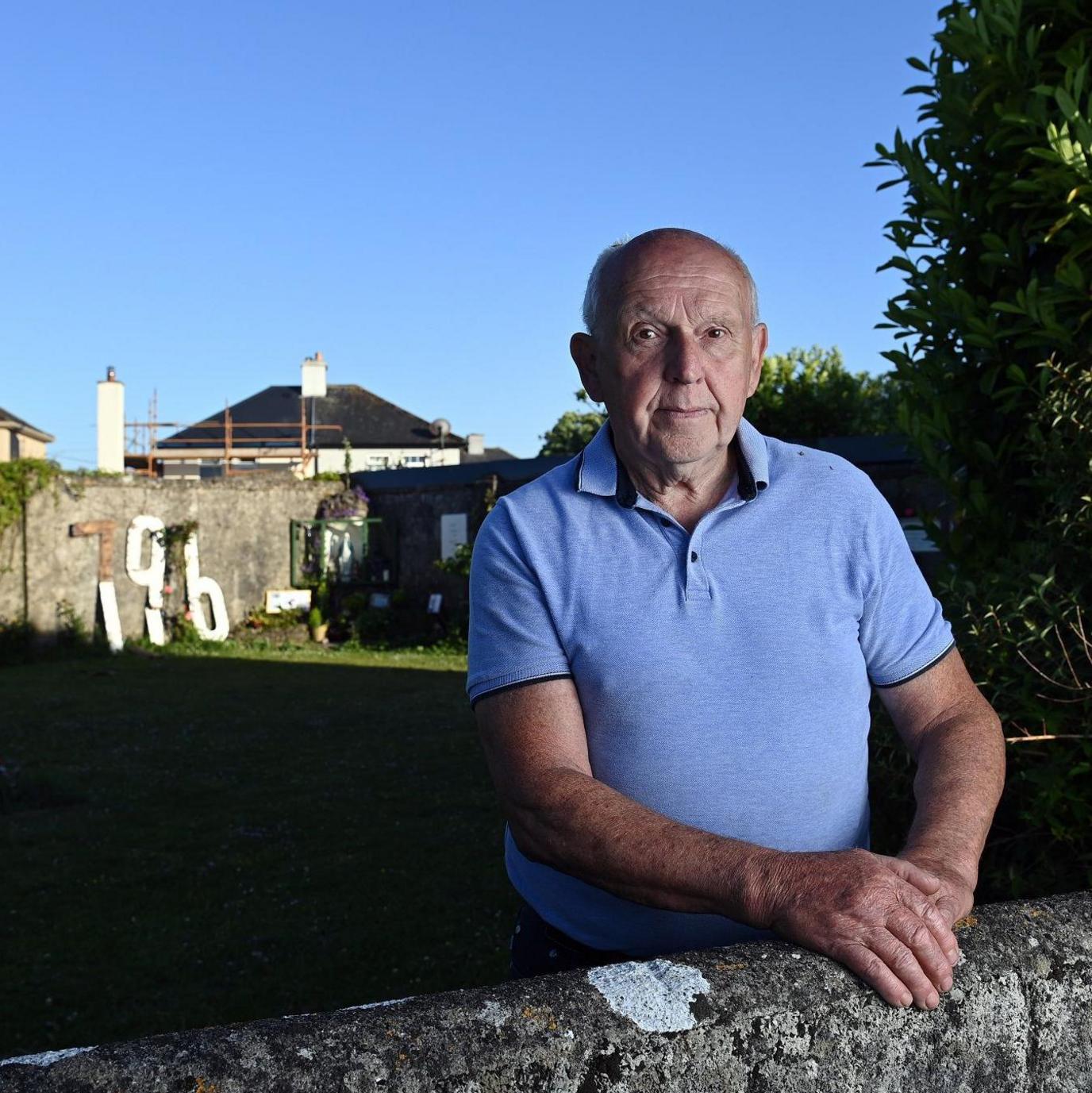A man in a blue polo shirt stands at a waist-high stone wall. In the background is a patch of lawn that continues until a high boundary wall. Part of the wall is covered in ivy - on another part can be seen the number 796 in large, white wooden lettering. The man in the foreground is older, bald and has his hands placed on the low wall. It is day time and the sky is clear blue.