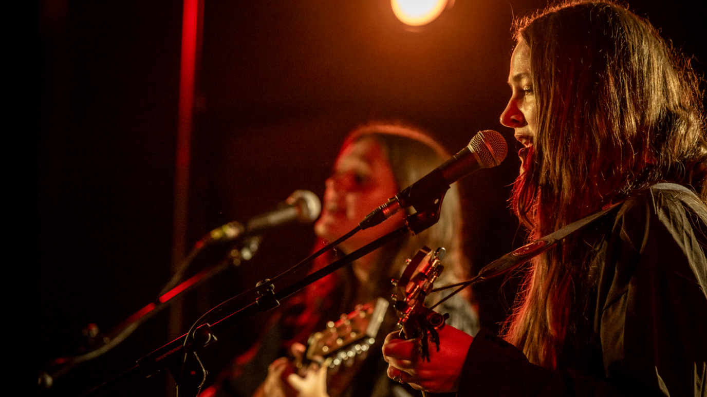 A side view of two members of the Staves singing and playing guitar with a black background