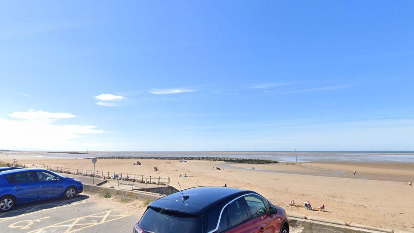 A stretch of beach is pictured in the background with people enjoying the sunshine in the distance, parked cars can be seen in the foreground and there are blue skies above