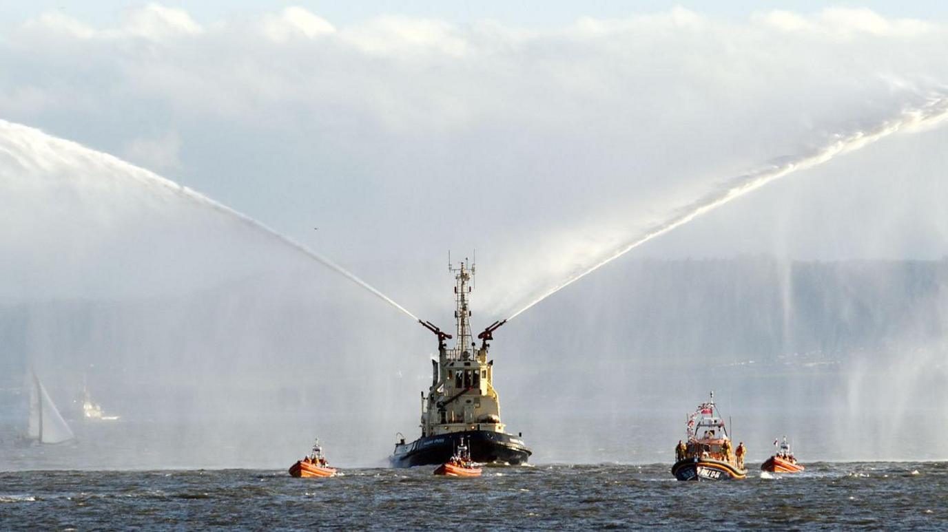 A large white and blue boat spraying water out of its sides. Smaller orange boats surround it.