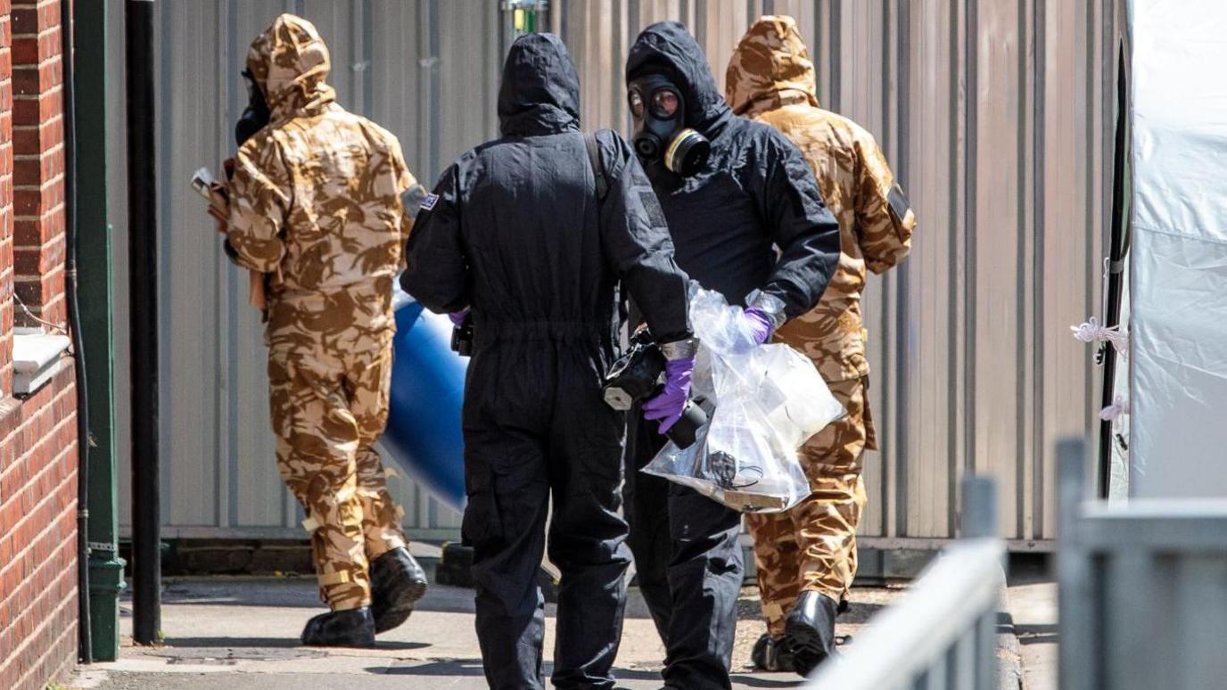 Members of the emergency services in protective gear while searching Dawn Sturgess home in Wiltshire. Two are in black full body suits and gas marks. Two are in camouflage coloured suits and face masks.