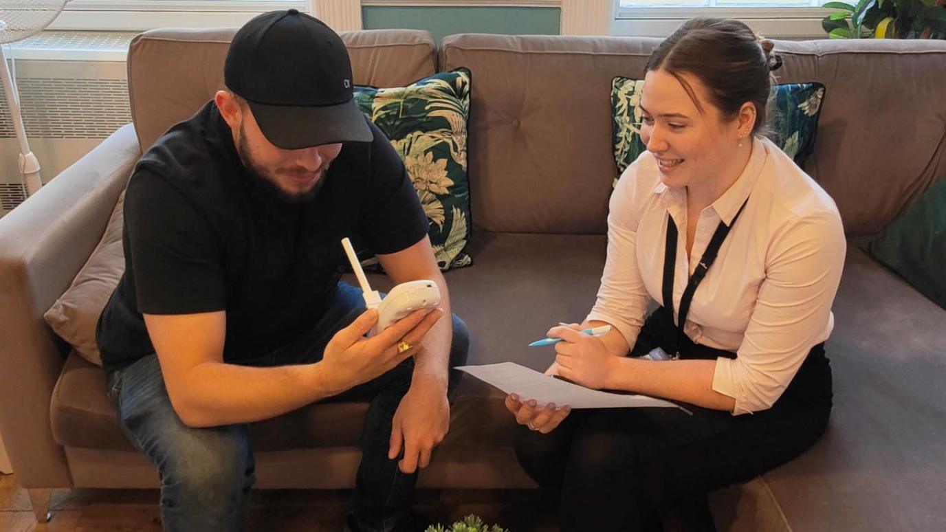 Man and woman sat together on a sofa. She is wearing a white shirt and writing things on a piece of paper. He is wearing a black top and cap while looking at a monitor to measure breath.