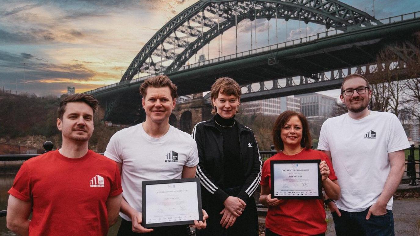 The Sunderland Music City Team – Marty Longstaff (left, in red Sunderland Music City T-shirt), Frankie Francis (centre left, in white Sunderland Music City T-shirt), Michelle Daurat (centre right, in red Sunderland Music City T-shirt) and Andrew Dipper (right, in white Sunderland Music City T-shirt) with managing director of the Music Cities Network, Lena Ingwersen (centre, in black Adidas tracksuit with three white stripes across the sleeves) and holding the membership certificate. Marty and Frankie have short brown hair, swept to the back. Michelle has shoulder-length straight brown hair with a side fringe. Andrew is wearing glasses and has a buzz cut. Lena has short wavy brown hair with a fringe. 
Sunderland's Wearmouth Bridge is in the background. It is a green arch structure. 