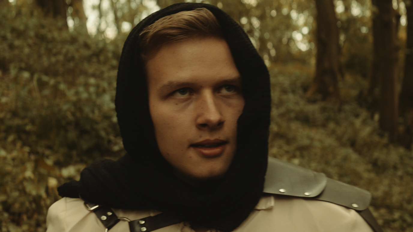 Ihor Kuraliesin, with a black scarf around his head and a blonde quiff of hair showing at the front. The forest is behind him and he is staring to his front left.