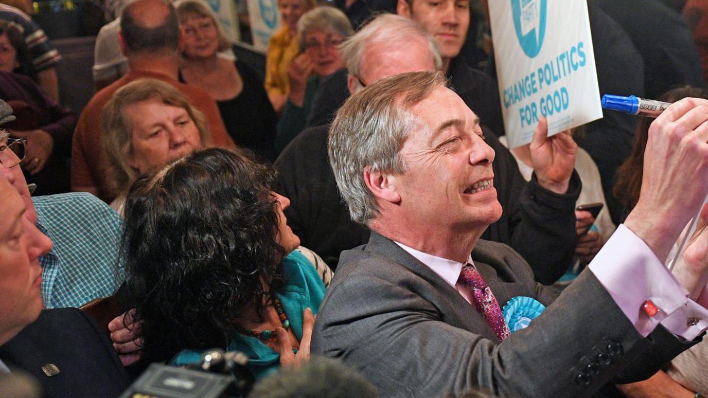 Nigel Farage, who has short greying hair and is wearing a suit, signs a placard for a supporter. He is inside Sugar Hut, which is packed full of people.