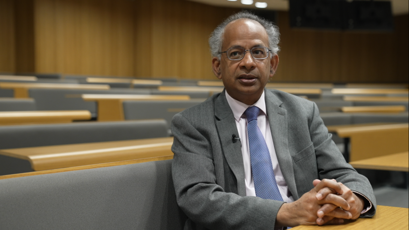 Professor Rama Thirunamachandran, wearing a grey suit, blue tie and white shirt. He is sitting in a lecture theatre with his hands clasped on the desk in front of him, looking to his right. 