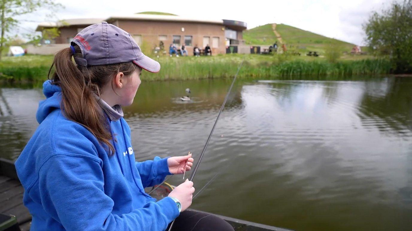 A young girl with long brown hair in a ponytail sites on a river bank with a fishing rod extended over a water way. She wears a faded purple cap and a blue hoodie