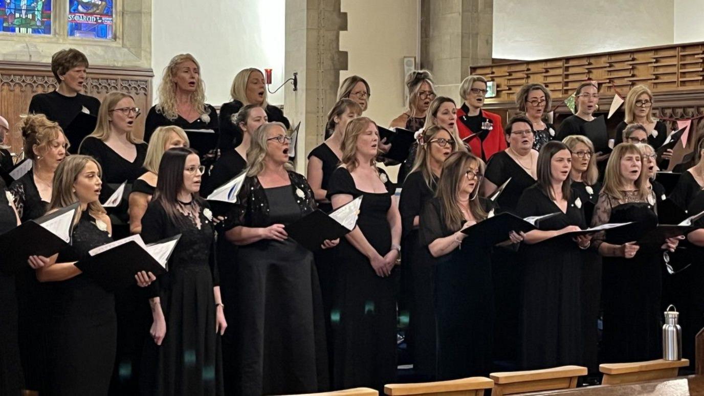 A choir is singing. Most are wearing black, and holding black folders with white pages in.