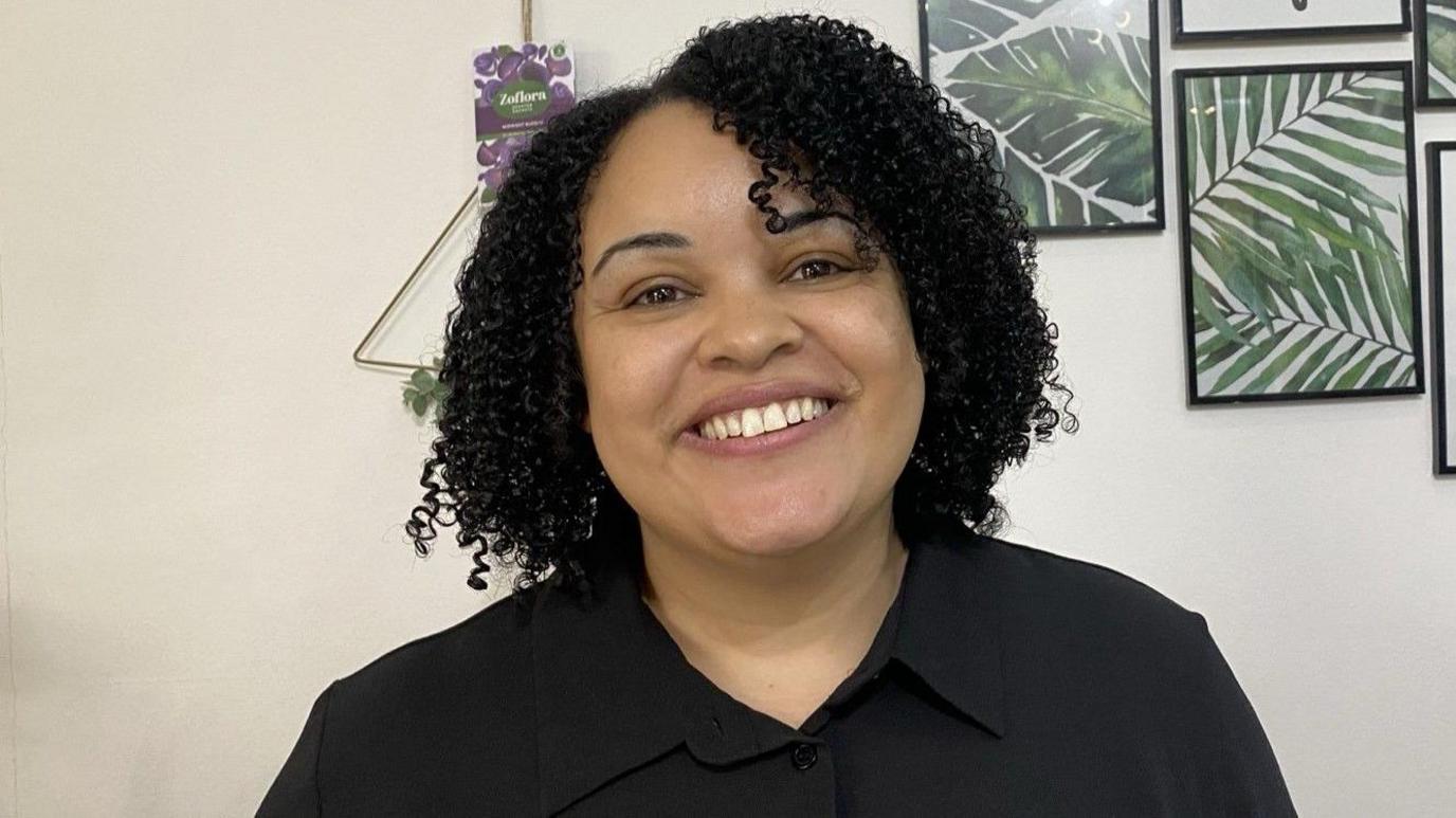 A woman with short black, curly hair to her shoulders smiles into the camera. She wears a black shirt and behind her are photos on the wall, with the ones visible showing the fronds of green ferns.