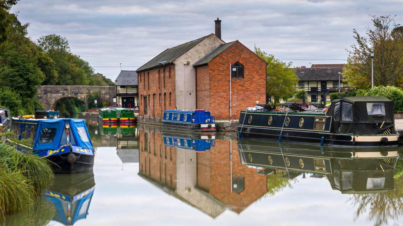 A view of the canal in Devizes showing boats and the Wharf Theatre, which is a red brick building. There is a small bridge over the canal and greenery on the river bank. 