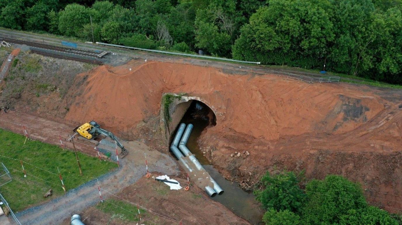 A aerial view of a railway line on top of an embankment with a tunnel running through it and metal pipes in the tunnel. There is also a yellow mechanical digger to the left of the tunnel