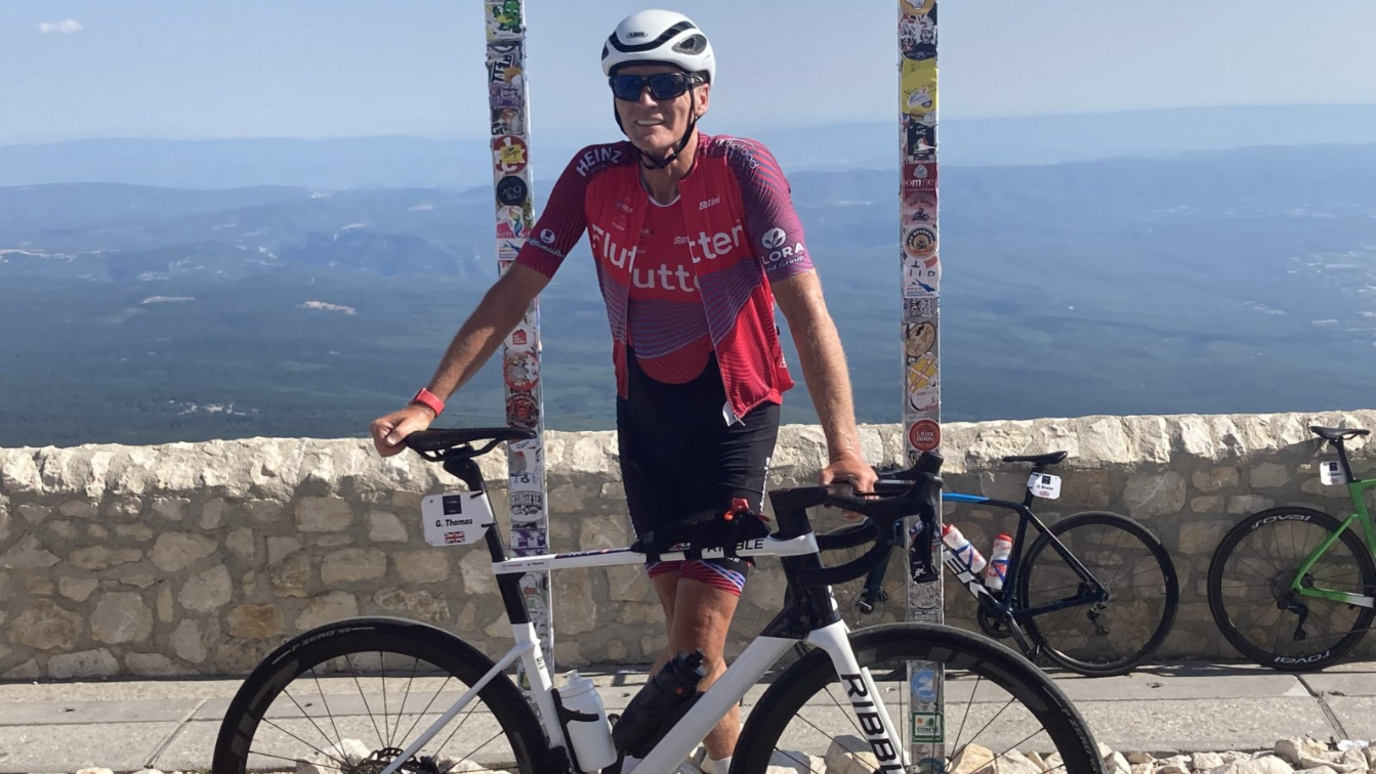 Thomas is stood in full cycling kit leaning on his bike by a low wall with the sea behind him.