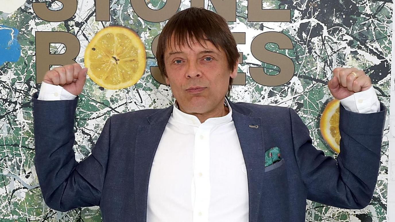 Mani with both arms raise standing in front of a large image of the Stone Roses' debut album artwork