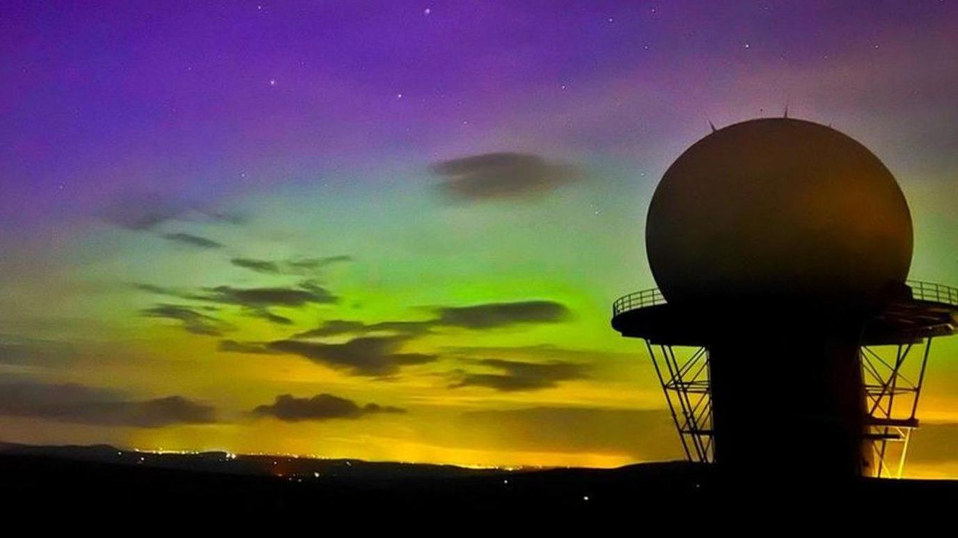 The structure is black or grey in the foreground. The ground is black and the sky features purple, green and yellow.