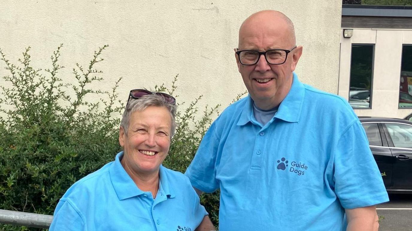 Anne Taylor and Steve Taylor stood together smiling wearing turquoise Guide Dogs polo shirts.